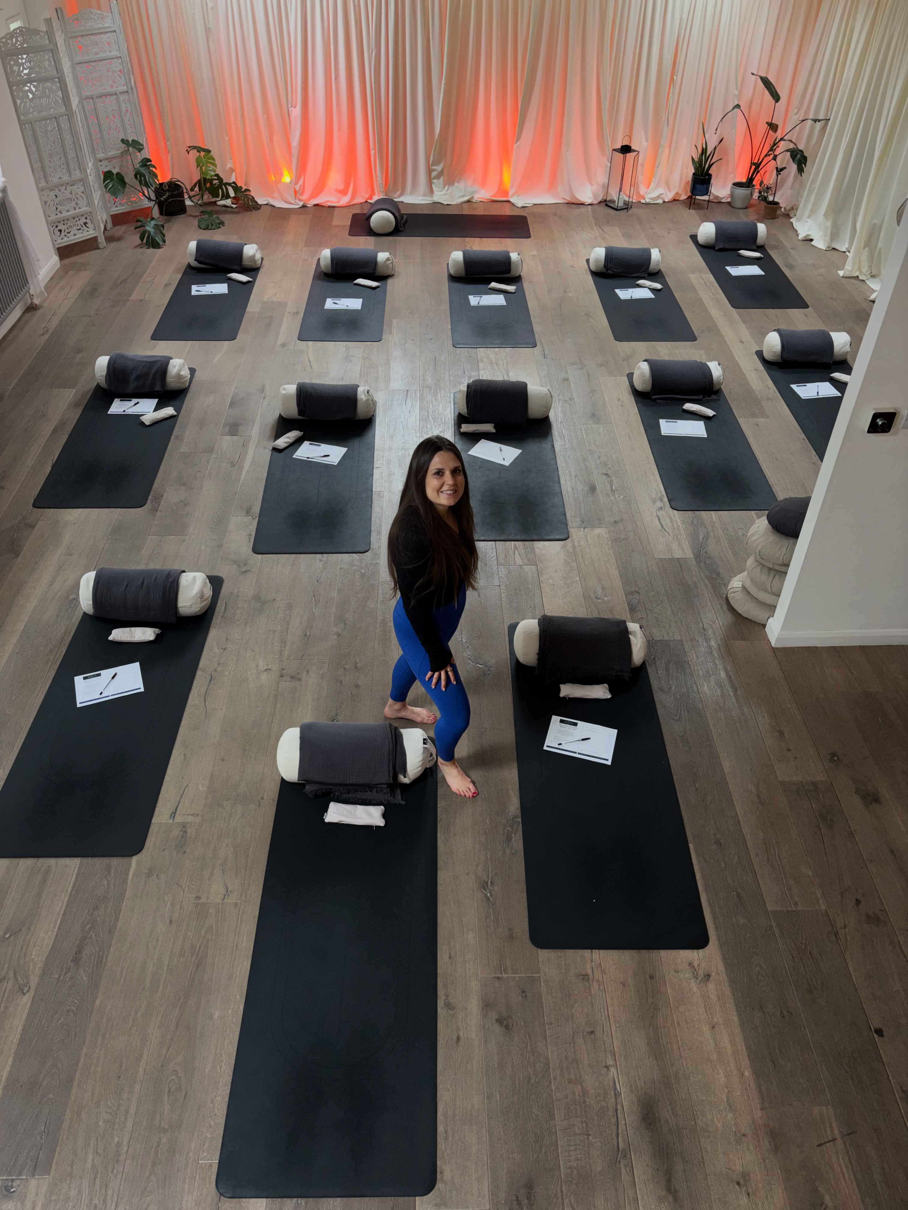 Lisa standing in the centre of a beautifully set-up wellness studio with yoga mats and warm coral lighting
