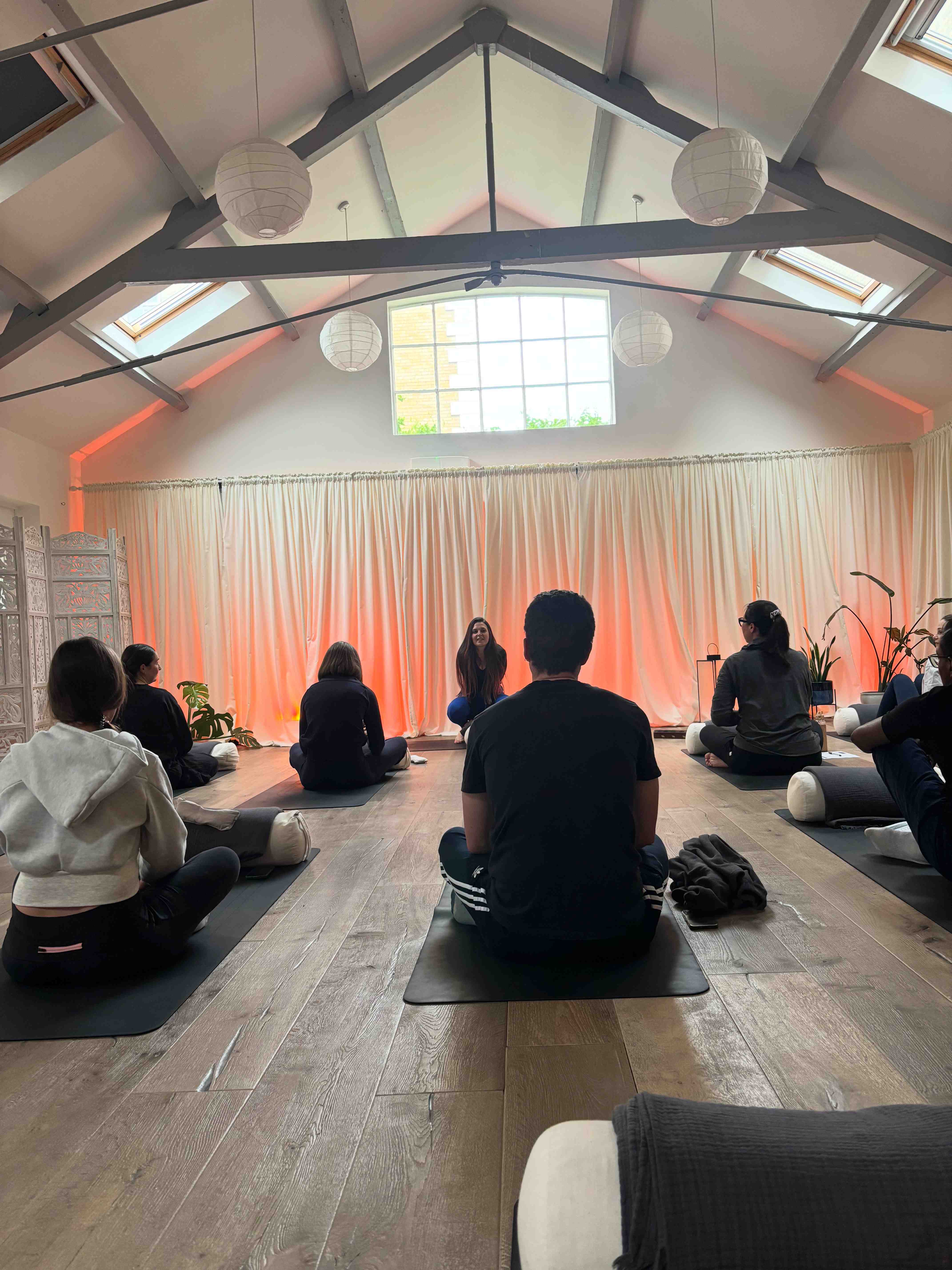 A group of people seated on yoga mats in a warm studio with paper lanterns, in a community wellbeing session