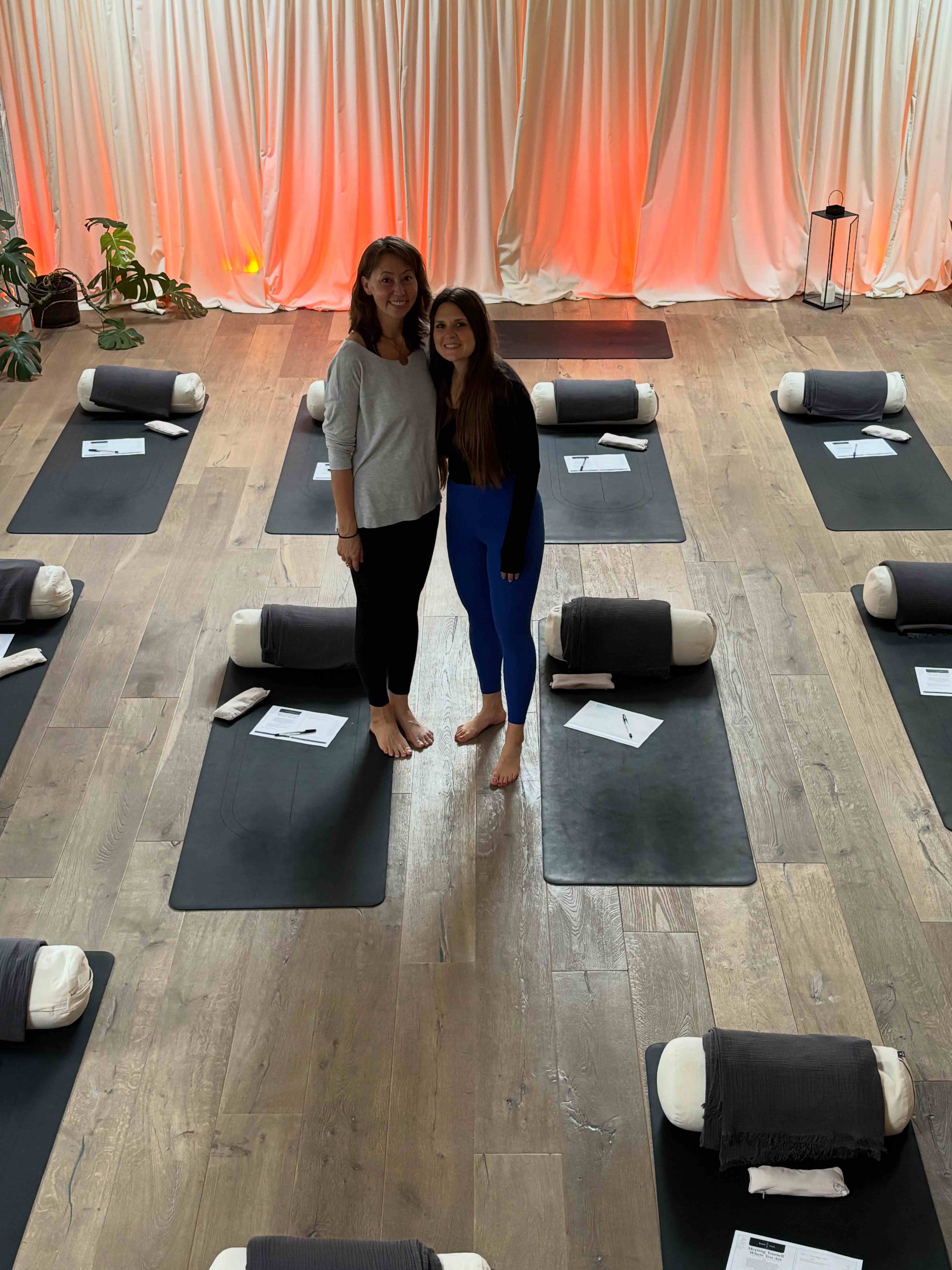 Two practitioners standing together in the Kindroot studio surrounded by yoga mats and bolsters